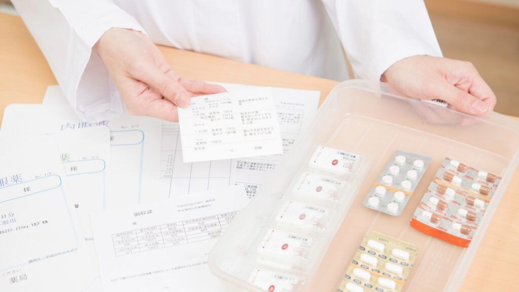デスク上に広げられた領収書や薬の説明書を確認しながら、トレイにまとめられた薬のシートを指で示す薬剤師。