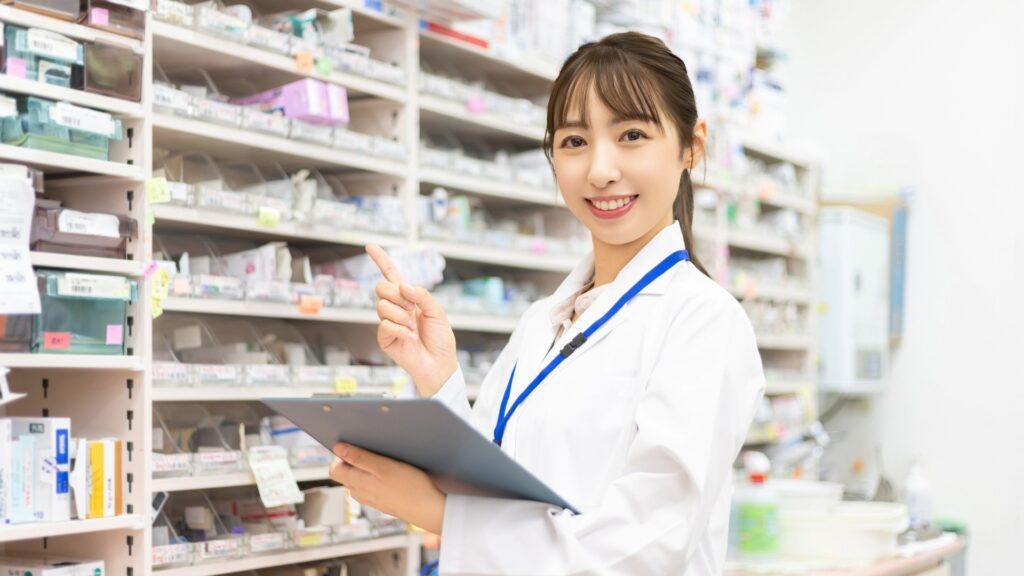 多くの薬が並ぶ棚の前で、白衣の女性がクリップボードを抱えながら、人差し指を立てて上を指している。